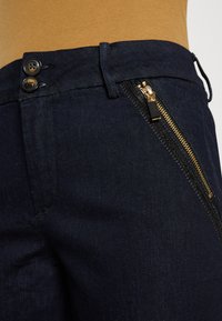 Jeans de mezclilla oscura con un zipper dorado en el lado izquierdo, dos botones en la cintura y una tela suave y ligeramente texturada.