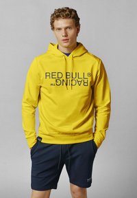 Geltona siuvinėta striukė su priekinėmis kišenėmis, ant kurios išdidžiai parašyta juodais rašmenimis 2D BULL RACING. Derinama su tamsiai mėlynomis šortais. Lygus paviršius, laisvas siluetas.