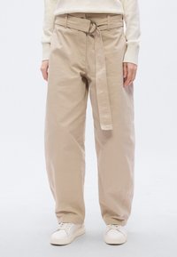 Μπεζ φαρδιά βαμβακερή παντελόνα με ψηλή μέση, φαρδιά μπατζάκια και ζώνη με κυκλικό ασημένιο πόρπη. Το σύνολο ολοκληρώνουν άσπρα αθλητικά παπούτσια.