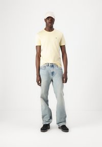 Tricou galben deschis, blugi lejeri de culoare albastră deschisă și adidași negri, asortați cu o șapcă de culoare deschisă; branding minim pe tricou.