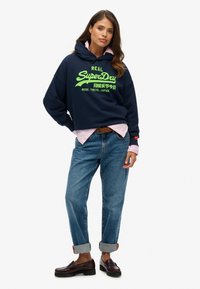 Sudadera navy con texto en verde neón, superpuesta a una camiseta rosa clara. Combinada con jeans claros remangados y mocasines oscuros. Diseño minimalista.