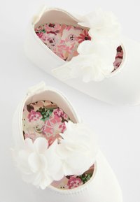 Weiße Stoffbabyschuhe mit floralen Verzierungen, runder Zehenform und gewellten Kanten. Das Innere zeigt ein mehrfarbiges Blumenmuster.