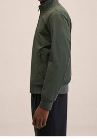 Zielona, lekka kurtka z wysokim kołnierzem, zamkniętymi bocznymi kieszeniami i elastycznymi mankietami, o gładkiej fakturze i dopasowanym kroju.