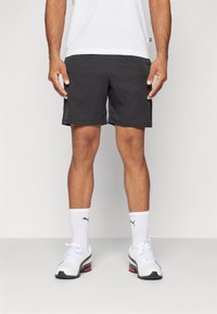 Hombre con pantalones cortos negros, calcetines deportivos blancos de Puma y zapatillas deportivas blancas, de pie frente a un fondo gris claro liso.