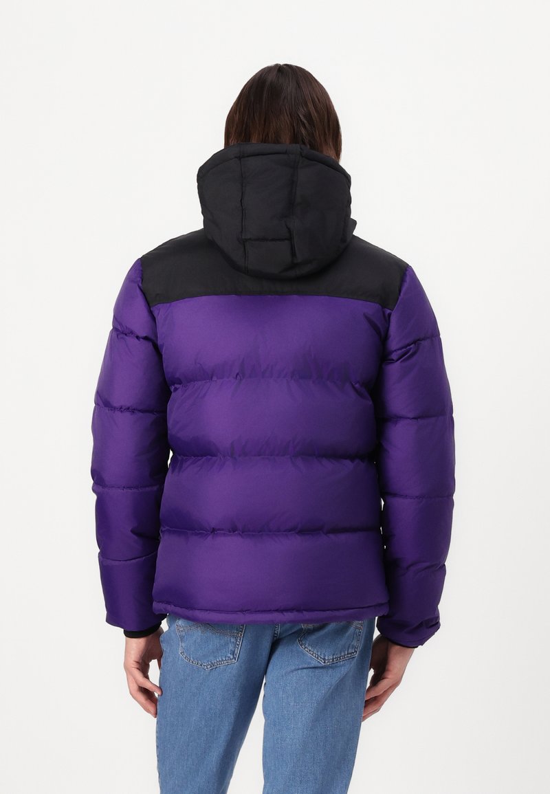 Giacca a vento viola con una sezione nera sulla spalla, dotata di cappuccio e texture trapuntata. La parte posteriore presenta un design aderente e polsini elasticizzati.