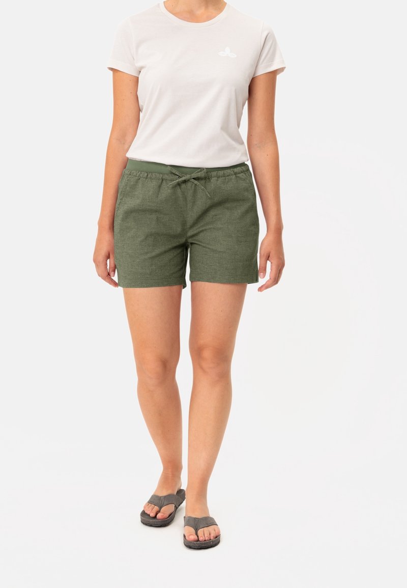 Femme portant une chemise blanche à manches courtes, un short vert olive avec cordon de serrage et des tongs grises, se tenant sur un fond blanc.
