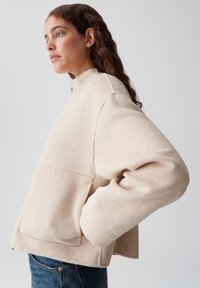 Maglione beige chiaro con colletto alto, maniche larghe e una grande tasca frontale. Tessuto morbido e dettagli di cucitura minimi.