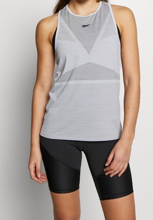 Frau trägt ein hellgraues, ärmelloses Sporttop mit Mesh-Details und schwarze, eng anliegende Radhosen, steht vor einer schlichten Wand.