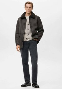 Giacca grigia con colletto foderato in pelliccia, abbinata a un maglione chiaro e jeans scuri. Il modello indossa scarpe nere, in piedi su uno sfondo semplice.