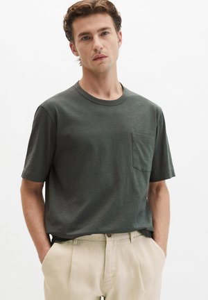 Jeune homme aux cheveux bouclés portant un t-shirt à manches courtes vert foncé avec une poche et un pantalon beige, debout les mains dans les poches.