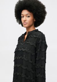 Jovem mulher com cabelo afro natural, vestindo uma blusa preta de manga longa com padrões texturizados em bordas onduladas, sobre um fundo claro.