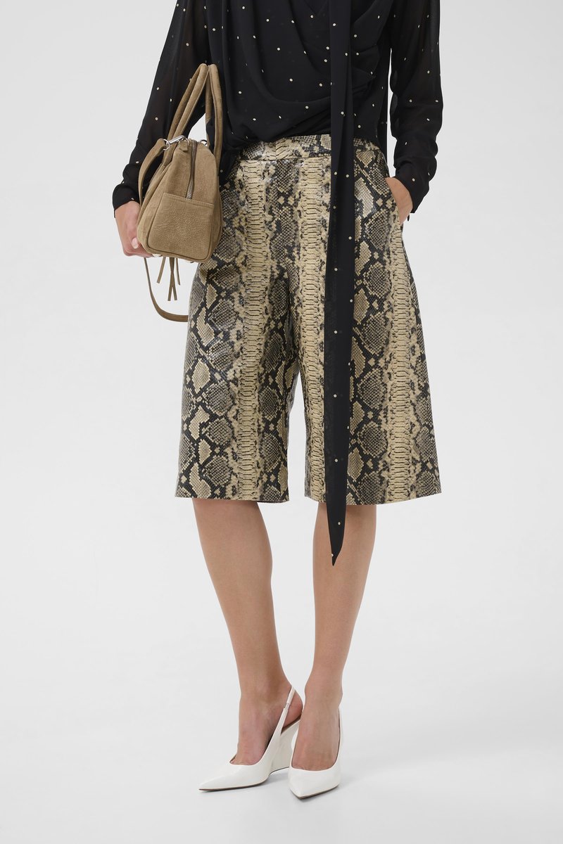 Γυναίκα που φοράει μπεζ και μαύρα σορτς μέχρι το γόνατο με σχέδιο φιδιού, μαύρο πουκάμισο με πουά, λευκές ψηλοτάκουνες γόβες με λουράκι και κρατά μια ταμπά τσάντα χειρός.