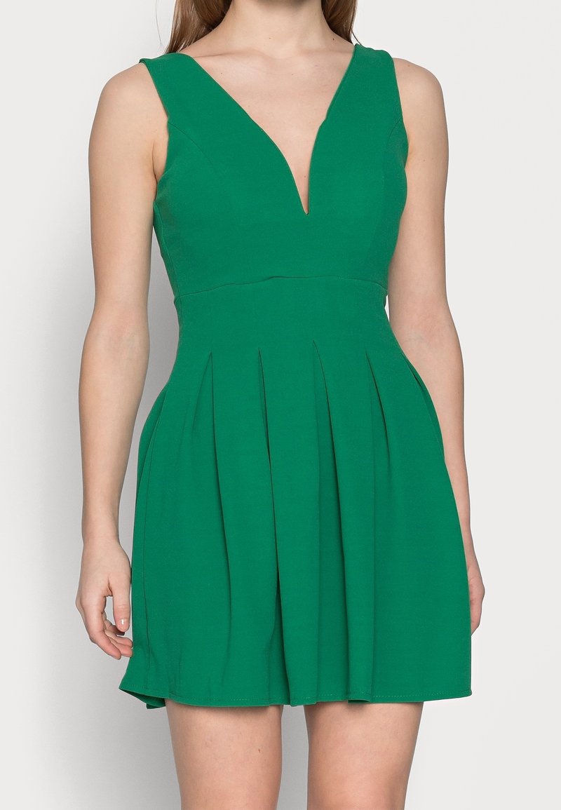 Grön ärmlös klänning med djup V-ringning, plisserad kjol, tillverkad av mjukt tyg, med strukturerade sömmar och en figurnära midja.