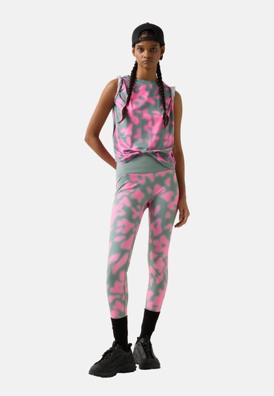 Jeune personne portant un haut sans manches gris et rose à motif et des leggings, casquette noire portée à l'envers, chaussettes noires et chaussures noires épaisses.