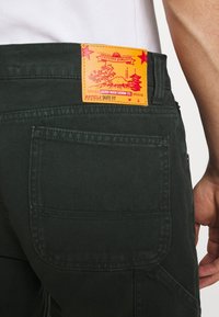 Tmavě zelené džínové kalhoty s texturovaným povrchem, s výraznou oranžovou koženou nášivkou na pásu s grafickými motivy a textem.