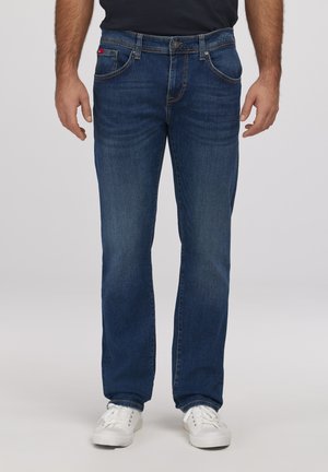 Mężczyzna w prostych niebieskich dżinsach i białych trampkach, stoi z rękami swobodnie opuszczonymi wzdłuż ciała na jednolitym tle.