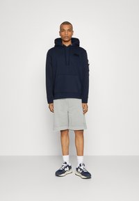 Hanorac bleumarin cu buzunar frontal și pantaloni scurți gri. Modelul poartă șosete albe și adidași bleumarin cu accente deschise. Design minimalist, croială casual.