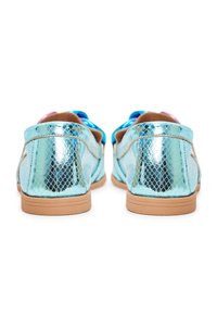 Mocassins à motif peau de serpent bleu métallique avec semelles marron clair vus de dos, sur fond blanc.