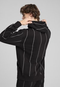 Crna dukserica s bijelim vertikalnim prugama, kapuljačom na užad i teksturiranim završetkom. Kroj je ležeran s rebrastim porubom.