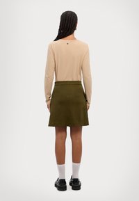 Lys beige langermet topp med logo-detalj på ryggen, kombinert med en mørk olivengrønn A-linjett skjørt med skjult glidelås og lommer.
