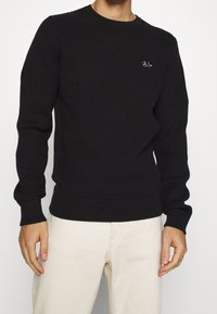 Maglione nero a coste con scollo rotondo, caratterizzato da un piccolo logo ricamato sul petto, completato da polsini e orlo aderenti.