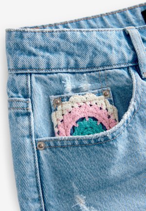 Los pantalones cortos de mezclilla azul claro presentan un parche decorativo de ganchillo en rosa, verde azulado y crema asomando por el bolsillo trasero. Detalles desgastados visibles.