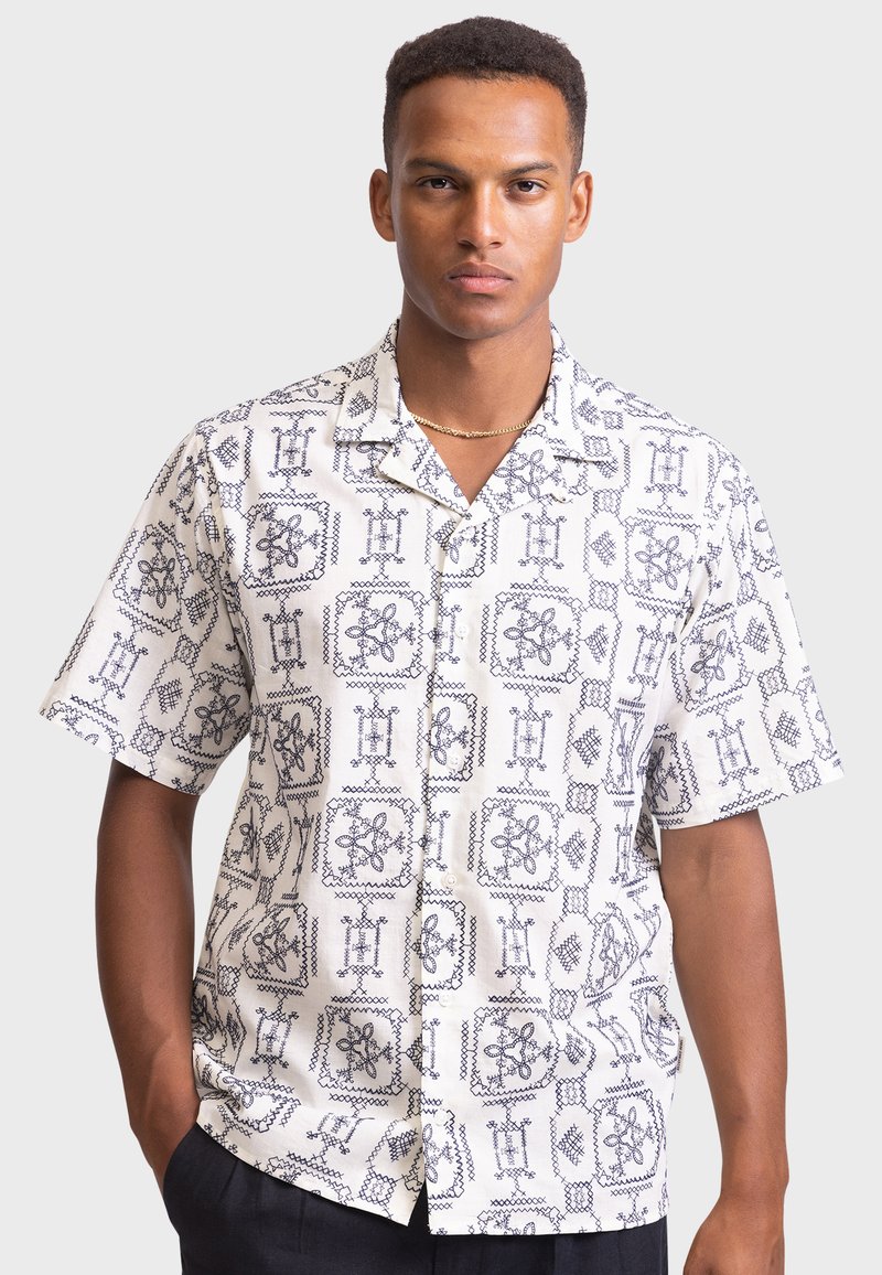 Short-sleeve button-up shirt in white fabric featuring a navy geometric pattern. Collar design, relaxed fit, and smooth texture are notable.