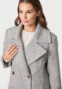 Cappotto grigio a spina di pesce con un ampio colletto, bottoni in legno doppi, e tessuto testurizzato, indossato sopra un top di colore chiaro.