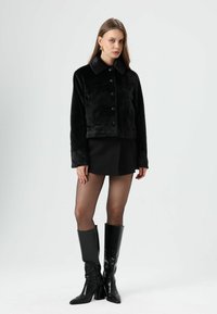 Veste en fausse fourrure noire avec col, fermeture à boutons sur le devant et deux poches latérales. Portée avec une jupe noire et assortie à des bottes noires montantes jusqu'aux genoux.