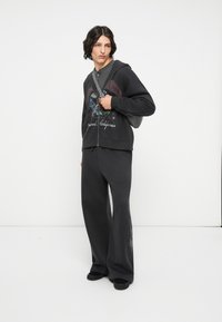 Felpa nera con zip e stampa grafica, abbinata a pantaloni della tuta neri e larghi con un delicato motivo. Modella con borsa a tracolla.