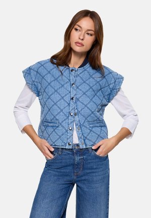 Mujer con chaleco acolchado azul abotonado sobre camisa blanca de manga larga, con las manos en los bolsillos, mirando hacia un lado contra un fondo liso.