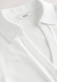 Polo de tela acanalada blanca con cuello doblado y etiqueta visible de la marca "NEXT" en el escote.