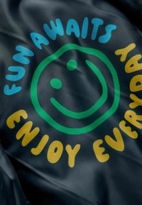 Tissu noir avec un visage souriant vert à l'intérieur d'un design circulaire, accompagnés du texte "LE PLAISIR VOUS ATTEND" en bleu et "PROFITEZ CHAQUE JOUR" en jaune.