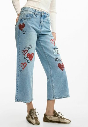 Jean bleu clair à jambes larges avec des motifs de cœurs rouges et noirs et un petit patch représentant un visage de dessin animé, porté avec des baskets beiges et un haut blanc.