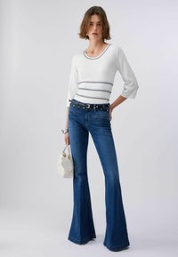 Femme portant un haut blanc ajusté avec des bordures noires, un jean bleu évasé avec une double ceinture noire, tenant un sac à main blanc, debout devant un fond uni.