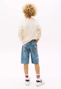 Maglione intrecciato beige chiaro, pantaloni shorts in denim blu, sneakers bianche con accenti blu navy e calzini a righe rossi e bianchi visibili alla caviglia.