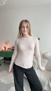 Világos rózsaszín, hosszú ujjú kötött pulóver, textúrált hullám mintával. Fekete, bő szárú nadrággal párosítva, egy világos beltéri térben.