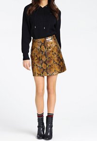 Donna in felpa nera, gonna mini in pelle di serpente lucida marrone con motivo, stivaletti neri e calzini neri con marchio su sfondo bianco.