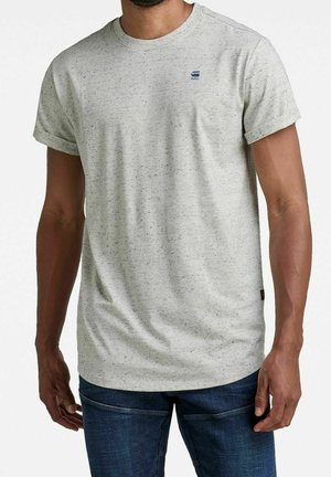 Mann trägt ein hellgraues, gesprenkeltes T-Shirt mit kurzen Ärmeln, die hochgekrempelt sind, und dunkelblaue Jeans, steht mit entspannten Armen.