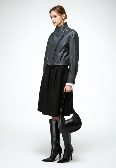 Vrouw die een zwarte leren cropped jas, een zwarte midirok, zwarte kniehoge laarzen met hoge hakken draagt en een zwarte leren handtas vasthoudt.