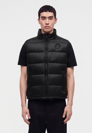 Jeune homme regardant devant lui, portant une doudoune noire sans manches par-dessus un t-shirt noir à manches courtes, sur un fond clair uni.