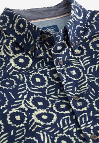 Námořnická košile na knoflíky s květinovým vzorem v světle žluté barvě. Materiál vypadá texturovaně, má klasický límec a hnědé knoflíky.