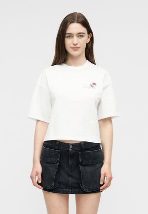 Mujer con cabello largo y castaño, lleva una camiseta blanca corta con un pequeño logo floral y una minifalda negra de mezclilla con grandes solapas en los bolsillos.