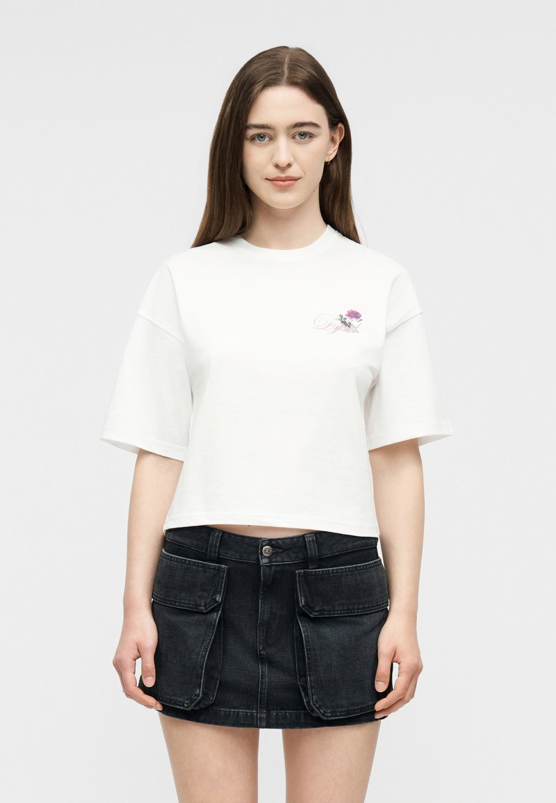 Γυναίκα με μακριά καστανά μαλλιά, φορώντας ένα λευκό κοντό T-shirt με μικρό λουλουδάτο λογότυπο και μια μαύρη μίνι φούστα από τζιν με μεγάλες τσέπες με καπάκια.