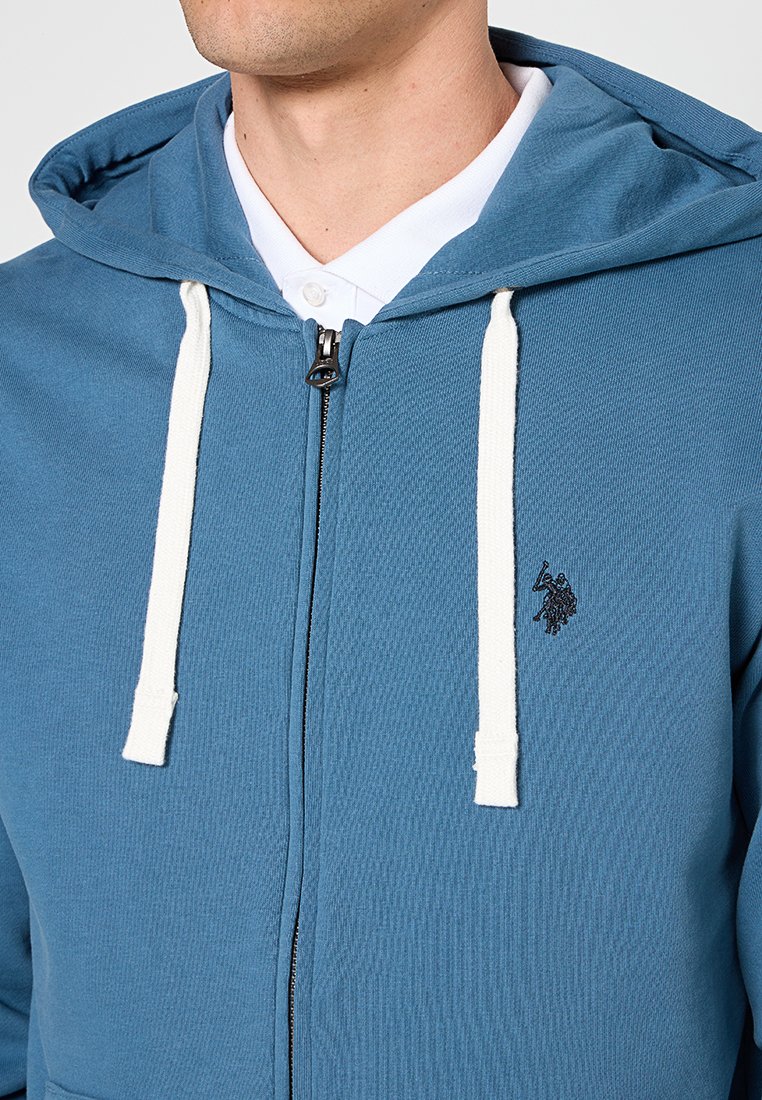 Mężczyzna noszący niebieską bluzę z kapturem na zamek z białymi sznurkami i małym czarnym haftowanym logiem, na białej koszuli z kołnierzykiem.