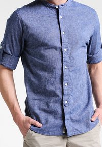 Mand iført en blå struktureret skjorte med oprullede ærmer og beige bukser, med hænderne i lommerne, stående foran en ensfarvet baggrund.