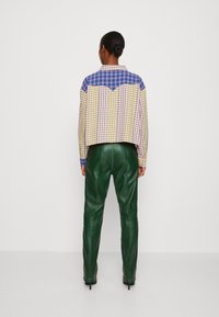 Hemd mit Knöpfen, blauem karriertem Einsatz, gelb-rosa kariertem Muster, kombiniert mit grünen Lederhosen und schwarzen High Heels.