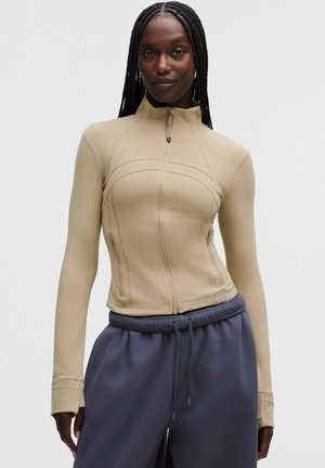 Vrouw met lange vlechten die een nauwsluitend beige vest met rits en losse donkergrijze joggingbroek draagt tegen een effen achtergrond.