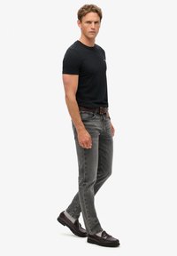 Camiseta negra de cuello redondo, jeans de denim gris con un acabado descolorido, cinturón de cuero marrón y mocasines oscuros. El modelo está en una pose neutral.
