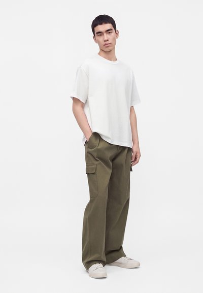 Jeune homme debout portant un t-shirt blanc uni, un pantalon cargo vert olive ample et des baskets blanches sur un fond blanc uni.